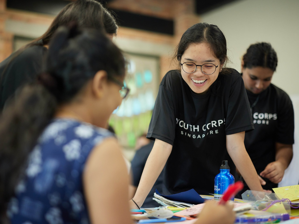 Several people in black Youth Corps Singapore shirts working at a table with craft supplies.